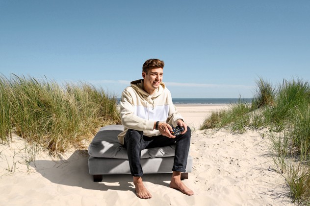 Junger Mann mit Controler in der Hand auf Hocker zwischen Dünen am Meer.